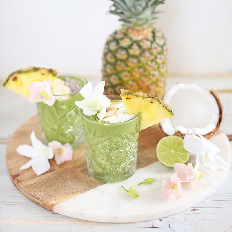 two small clear glasses filled with vegan green tropical smoothies garnished with slices of pineapple with additional pineapple and lime in the background