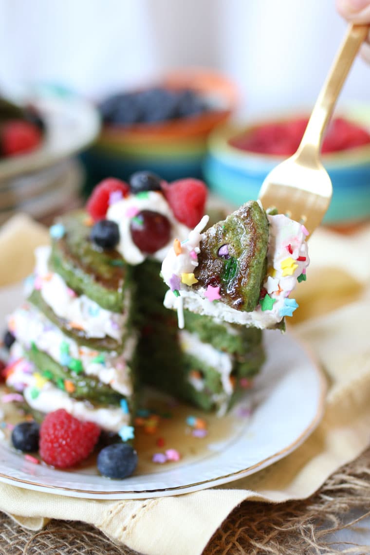 close up of a silver fork taking a bite out of a stack of vegan matcha rainbow pancakes garnished with fresh fruit