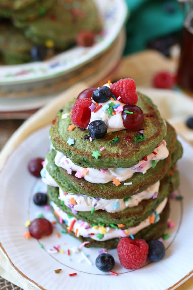 close up of a stack of vegan green st. patrick's day gluten free matcha rainbow pancakes garnished with sprinkles, fresh fruit, and whipped cream 