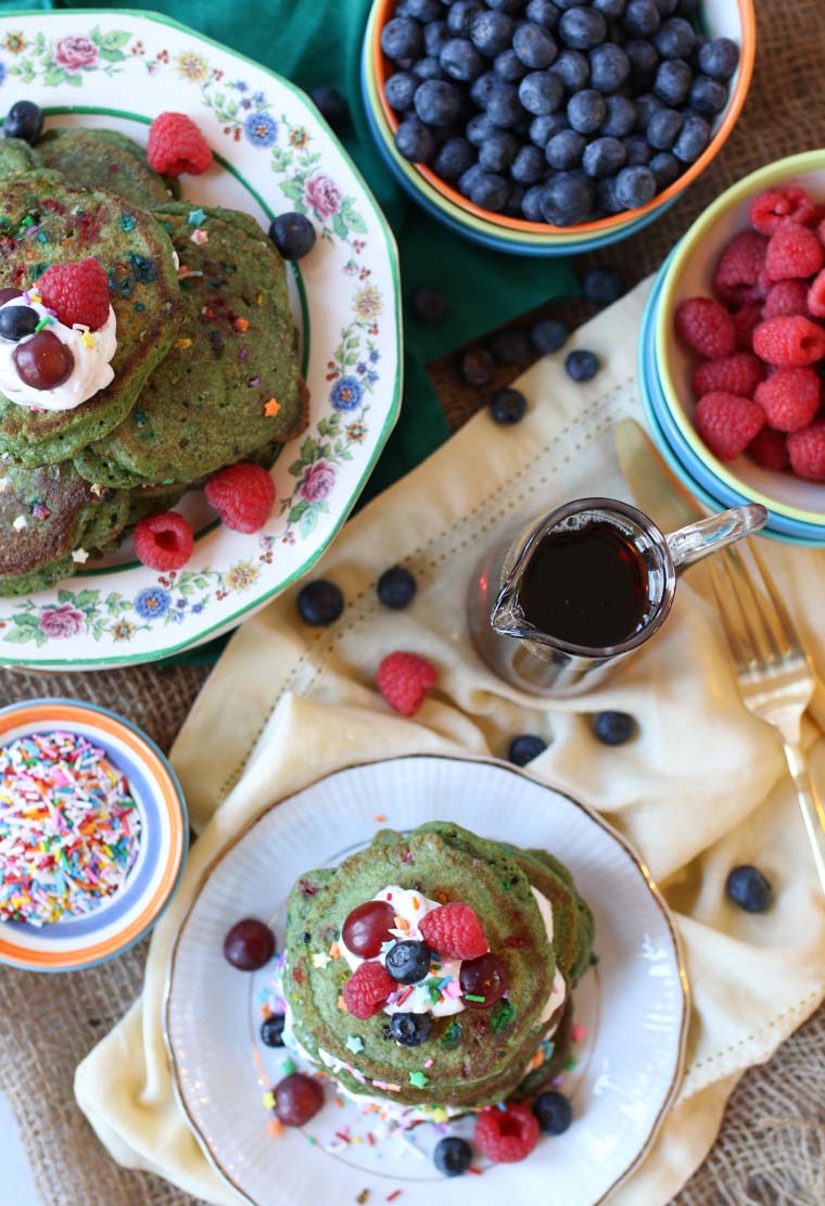A pile of matcha protein pancakes on a plate next to bowls of blueberries and raspberries.