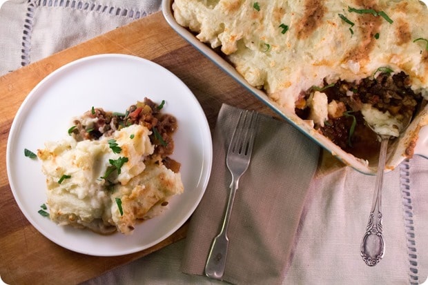 birds eye view of a piece of vegan and gluten free shepherd's pie garnished with fresh herbs on a white plate next to a silver fork and a casserole dish containing additional shepherd's pie 