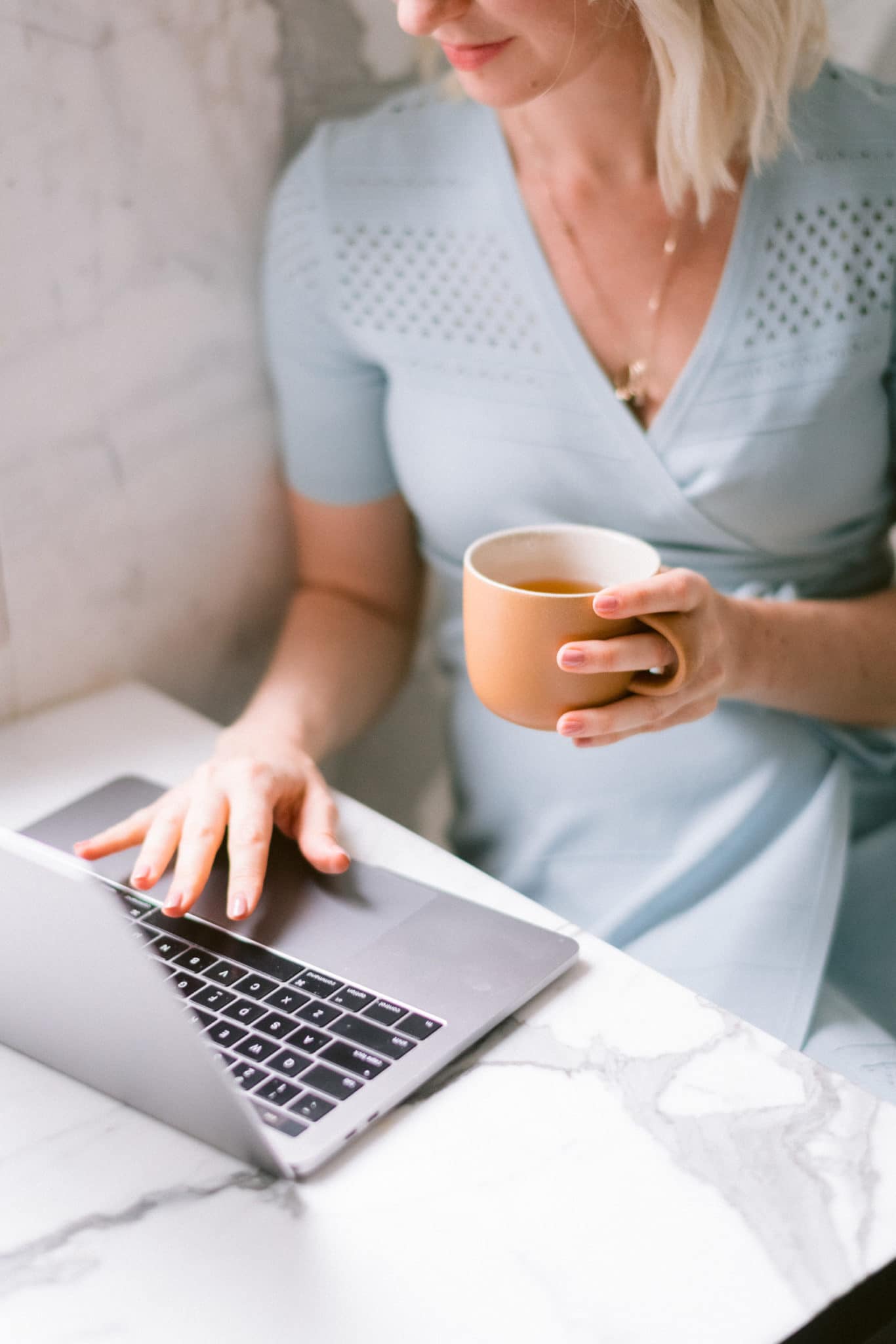 close up of abbey in blue dress with a coffee in her hand working from her laptop 