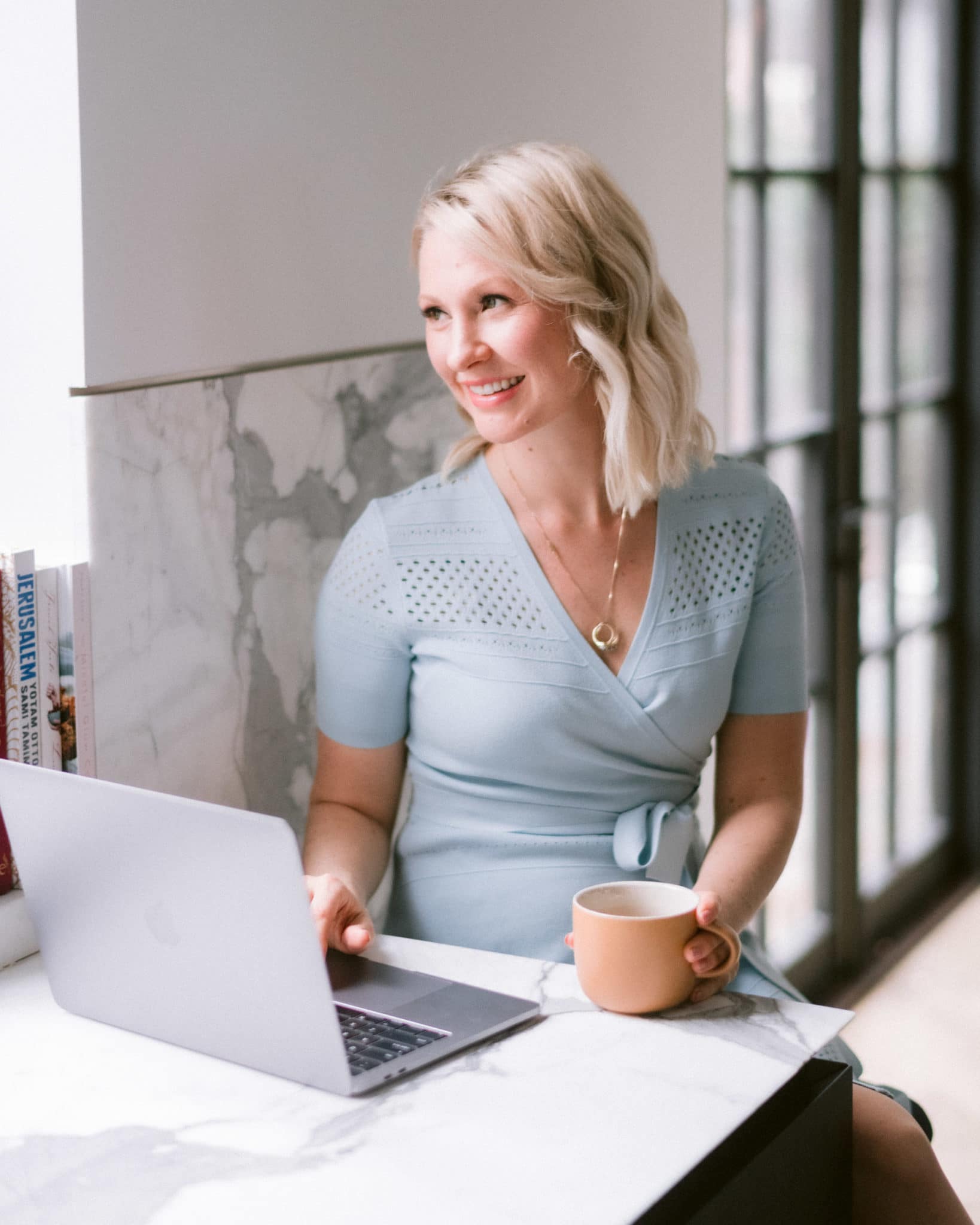 abbey in a blue dress with a brown mug in her hand working on her laptop 