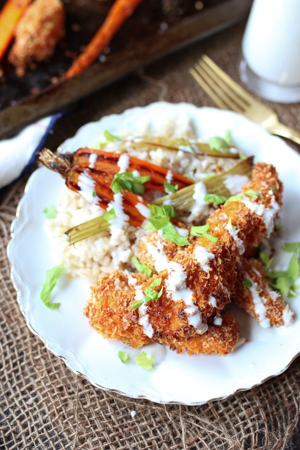 A white plate with buffalo chicken fingers, carrots, and grain with sauce drizzled on top.