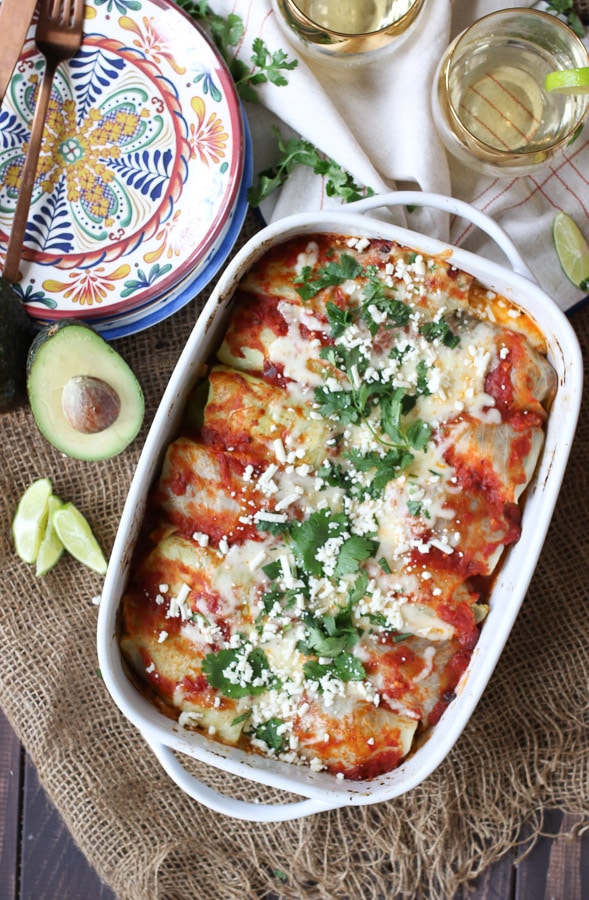 Cabbage roll enchiladas in a white casserole dish topped with cheese and cilantro next to an avocado halve, limes and plates and glasses.