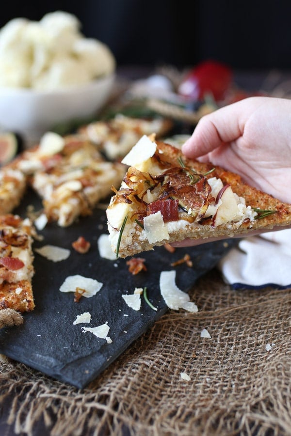 Hand holding a slice of apple, fig and onion cauliflower pizza flatbread.