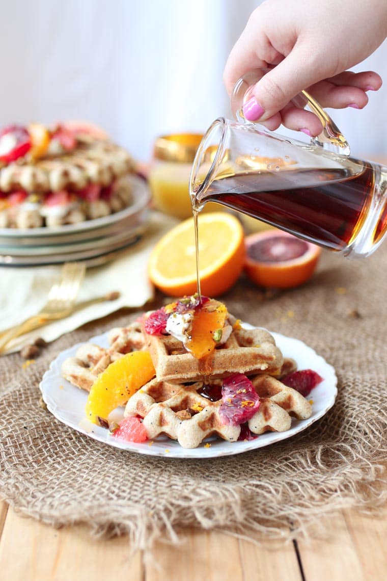 Hand pouring glass of maple syrup on a stack of vegan orange poppyseed waffles.