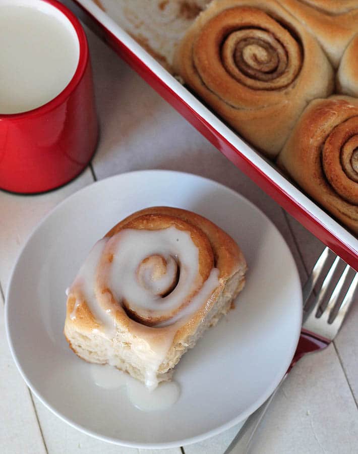 birds eye view of vegan cinnamon rolls on a white plate next to a baking dish filled with additional cinnamon rolls