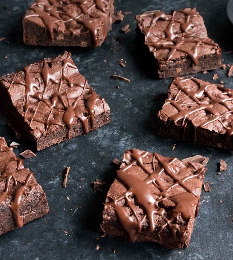 Multiple vegan brownies with chocolate drizzled on top on a dark backdrop.