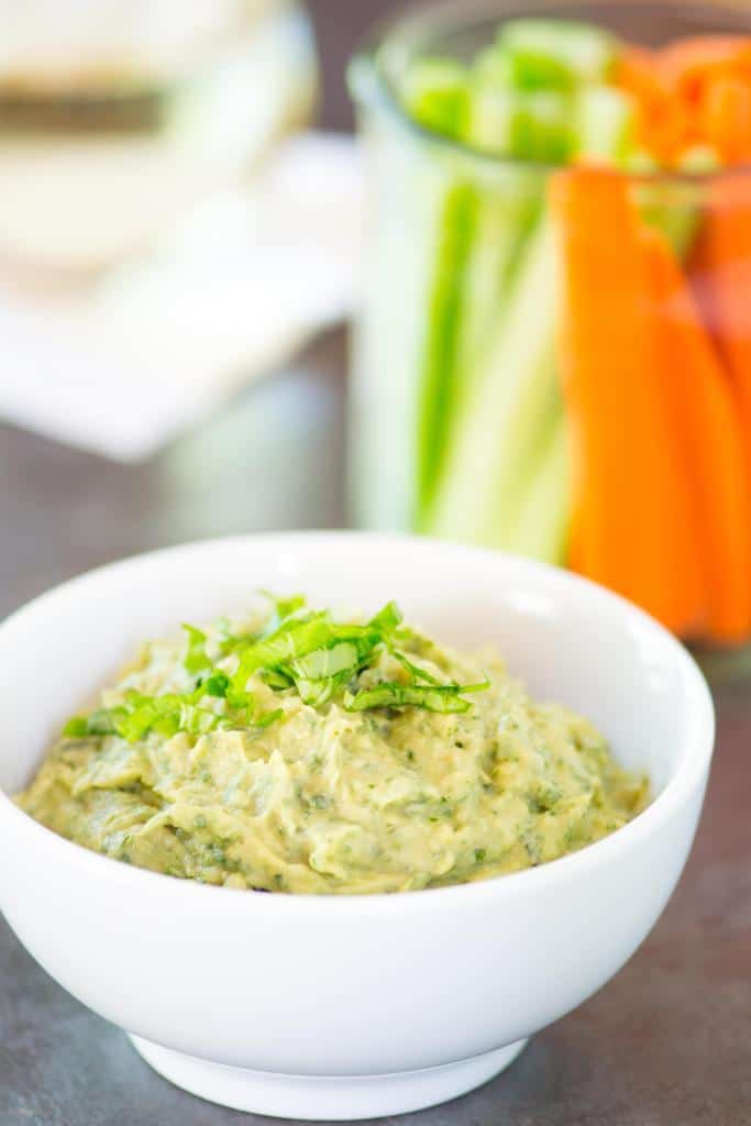 Close up image of vegan roasted garlic avocado and white bean dip garnished with fresh herbs inside of a white bowl, featuring slices carrots and cucumbers in a glass jar in the background