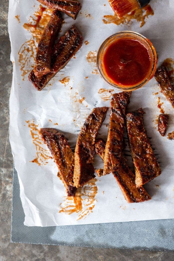 Birds eye view image of multiple vegan honey barbecue ribs served on top of a plate lined with parchment paper, and a small circular glass dish on the side containing additional honey barbecue sauce