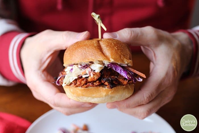 Close up image of two hands holding a vegan barbecue sandwich on a bun with coleslaw, held together by a small wooden skewer 