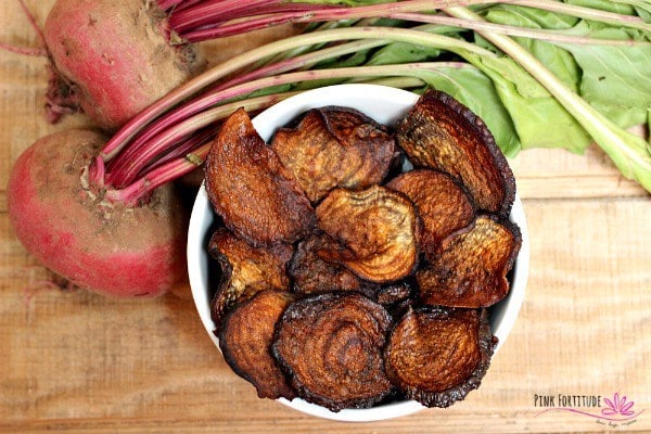 Birds eye image of vegan baked beet chips served in a large white bowl placed next to two raw beetroots, atop of a wooden surface