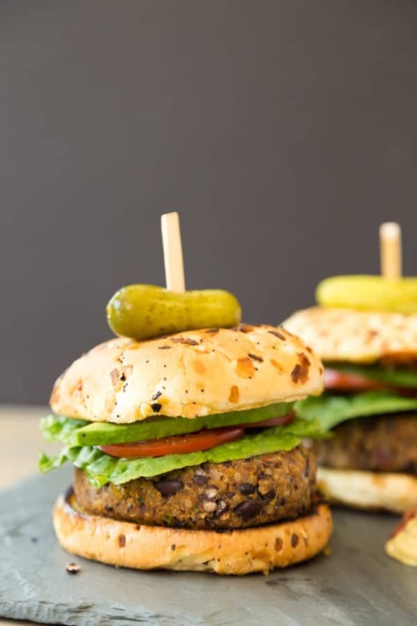 Close up image of two vegan spicy black bean and quinoa burgers inside hamburger buns, garnished with avocado, tomatoes, and lettuce, held together by a wooden skewer with a small pickle on the top