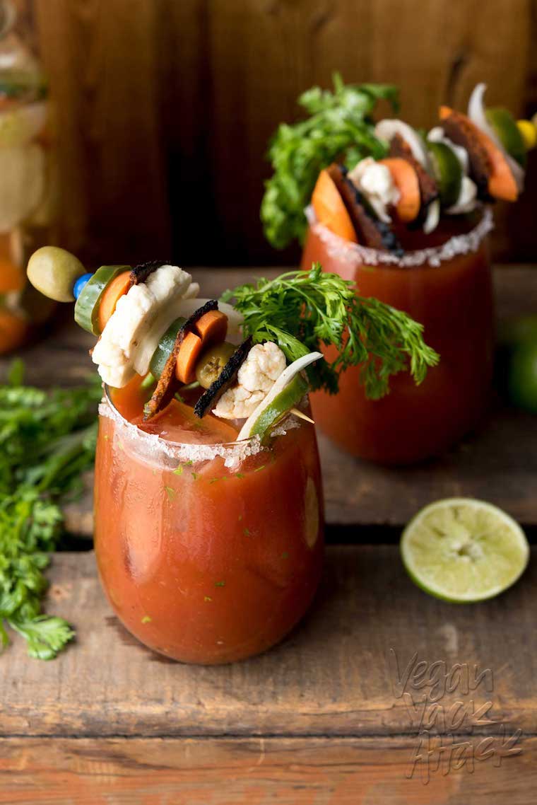 Medium close up shot of vegan loaded chipotle bloody mary in a clear glass garnished with loaded savoury skewer, fresh lime, and fresh herbs, on top of a wooden surface