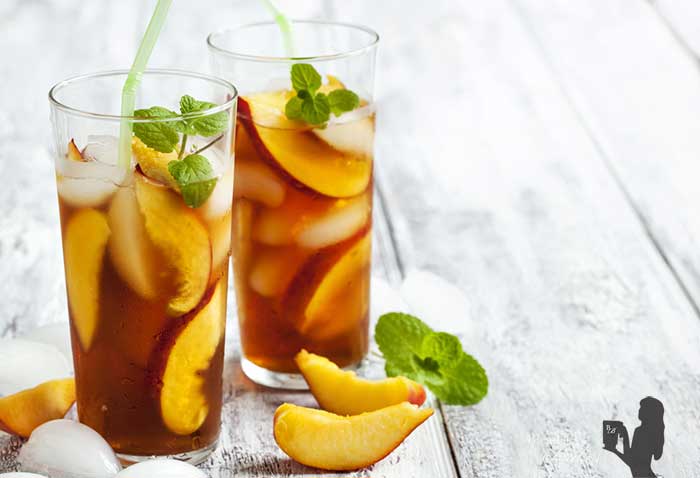 Medium close up image of two vegan peach limeade bourbon cocktails in tall clear glasses with green straws atop a wooden surface, garnished with fresh mint and additional fresh peaches