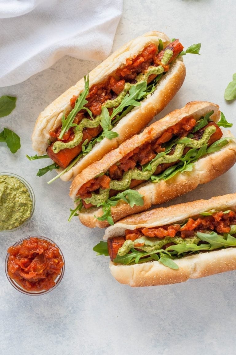 Birds eye view of three plant-based balsamic grilled carrot dogs inside hotdog buns, garnished with pesto, tomato jam, and greens, presented on a white marble serving dish, next to two small clear bowls containing additional garnishes