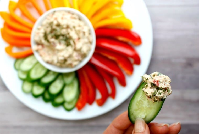 Birds eye view shot of a hand holding a slice of cucumber dipped into vegan chives and garlic cashew spread in the foreground, with additional sliced cucumbers, bell peppers, and carrots on a large white dish surrounding a white bowl containing the dip in the background