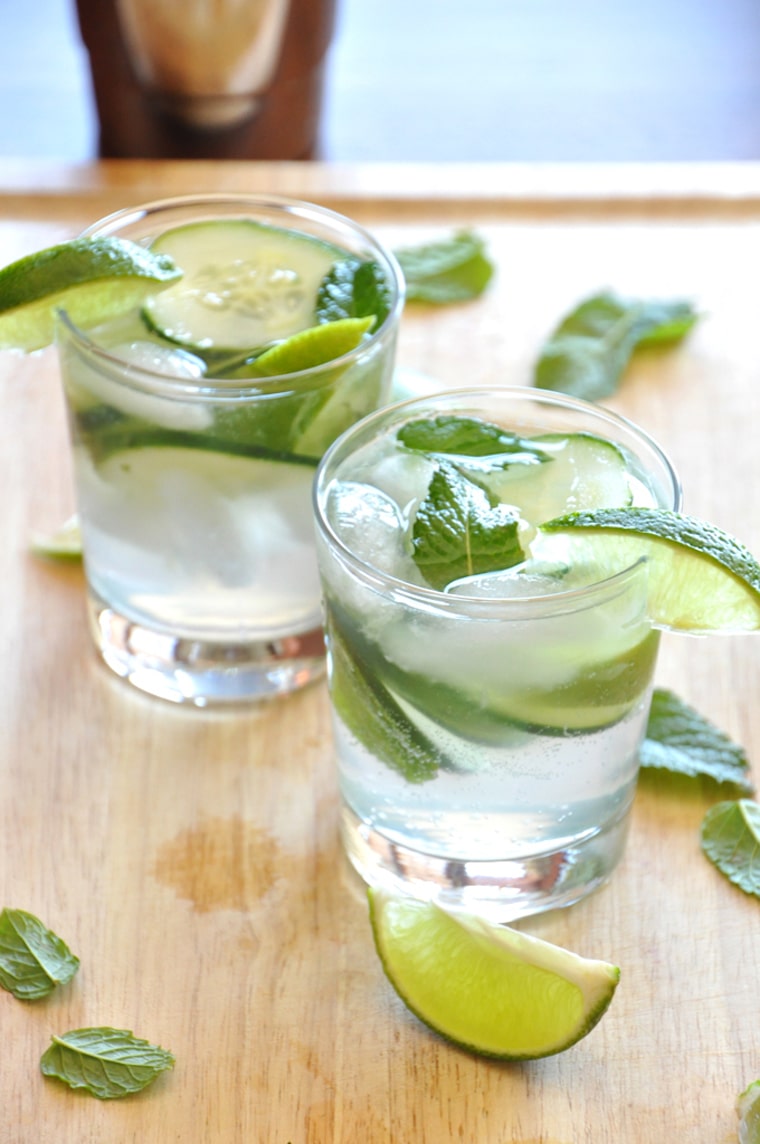 Close up image of two clear shot glasses filled with vegan cucumber cooler cocktails garnished with fresh mint and lime wedges atop a wooden surface