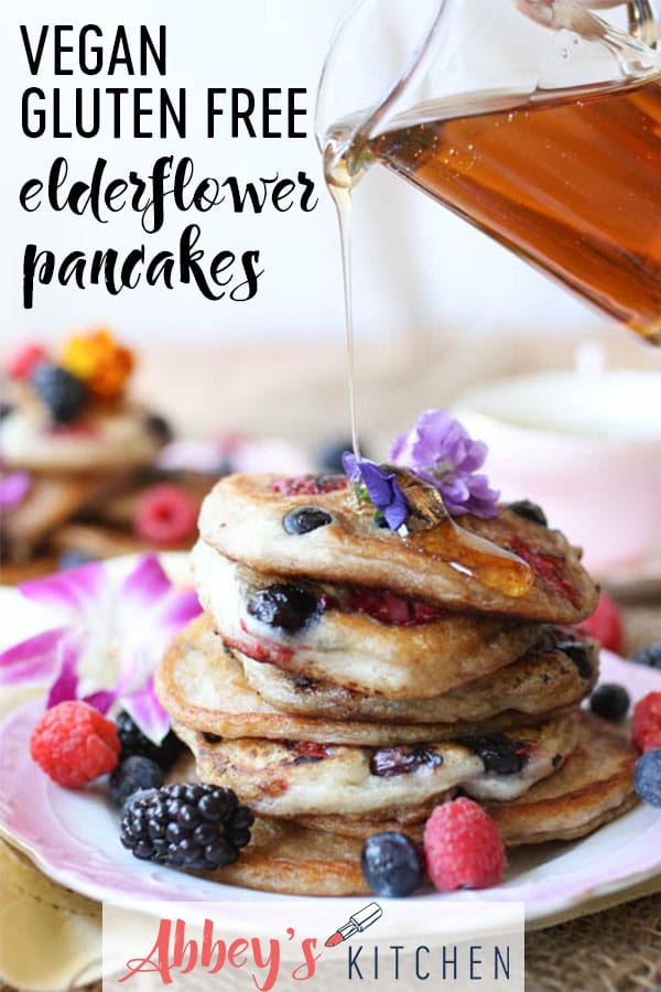 Pinterest image of an image of a plate of elderflower pancakes with berries and edible flowers around with syrup poured on with the text overlay \"vegan gluten free elder flower pancakes.\"