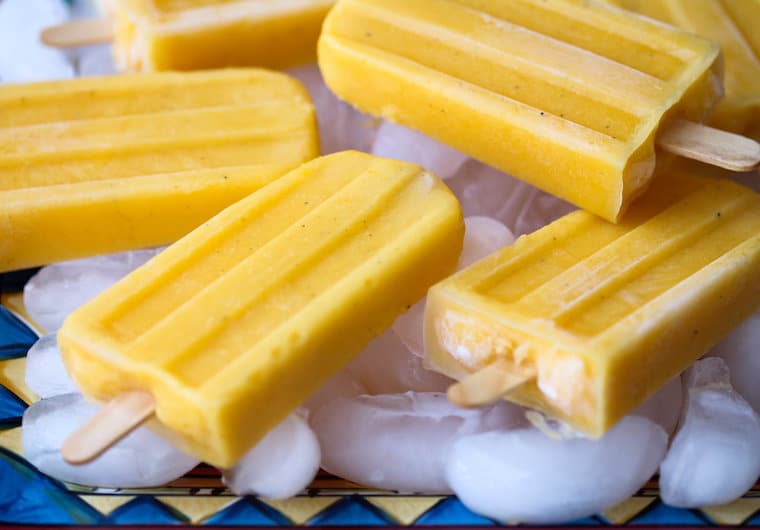 Close up of multiple vegan mango lassi popsicles with wooden popsicle sticks, laying on top of ice 
