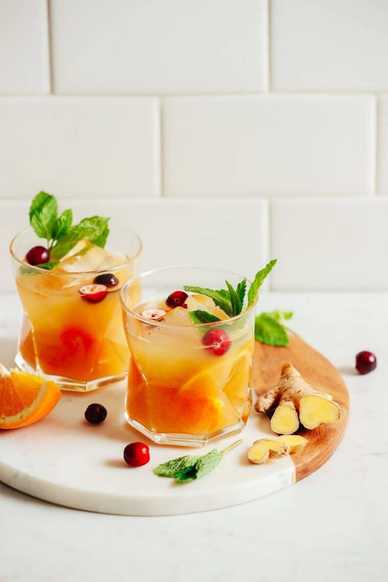 Close up of orange infused whiskey ginger in a clear glass against a white tile background, garnished with fresh cherries, mint, ginger, and orange slices 