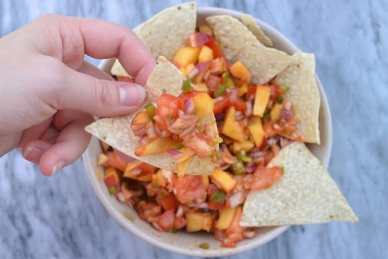 Close up image of a hand picking up a chip dipped in a bowl of vegan sweet and spicy peach jalapeno salsa against a white and grey marble background 