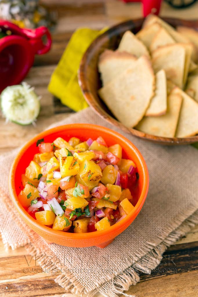 Close up image of vegan grilled pineapple salsa inside an orange bowl alongside a brown bowl containing tortilla chips, all sitting on top of placemats on a wooden table