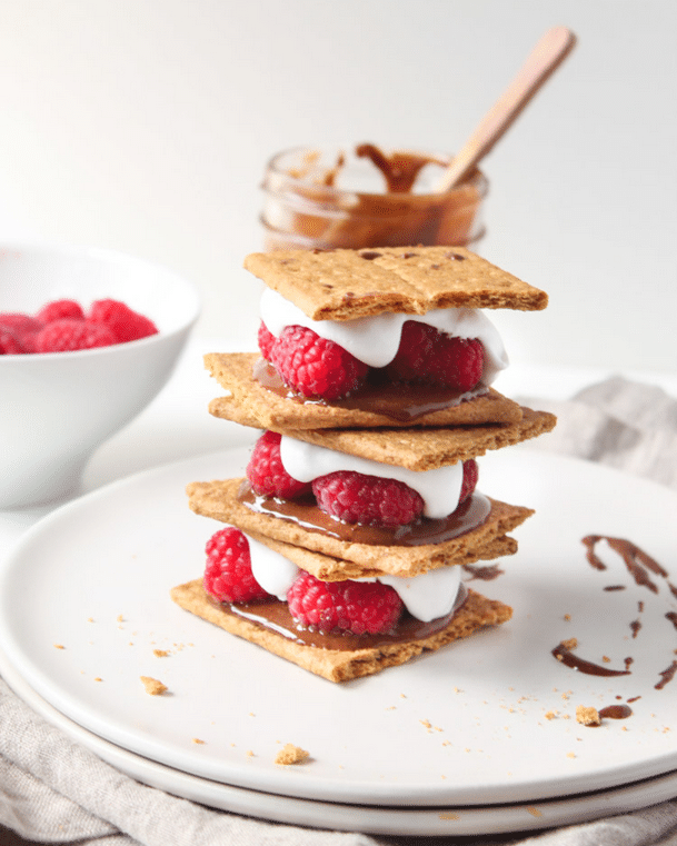 Vegan raspberry and coconut cream nutella smores stacked on top of one another on a circular white plate, garnished with additional nutella sauce, featuring a white bowl filled with raspberries and a glass jar of nutella in the background 