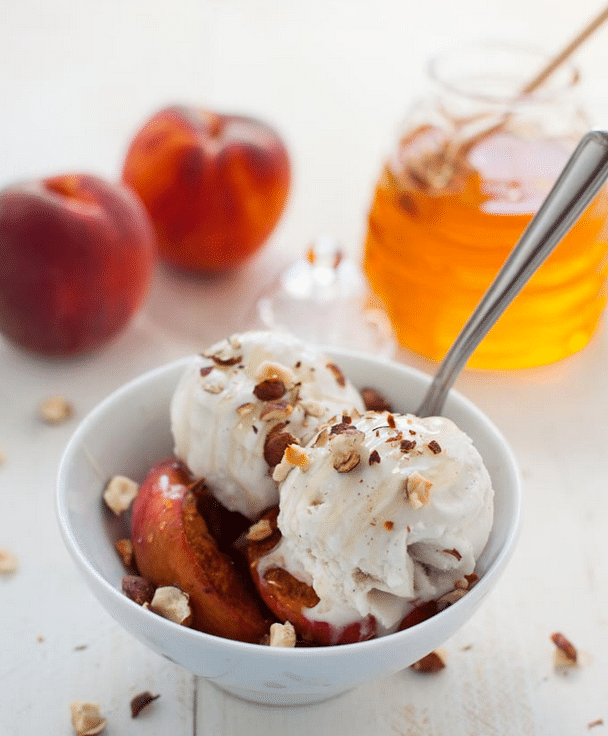 Plant-based roasted peach ice cream sunday served in a white bowl garnished with chopped nuts, and additional peaches and a jar of honey in the background