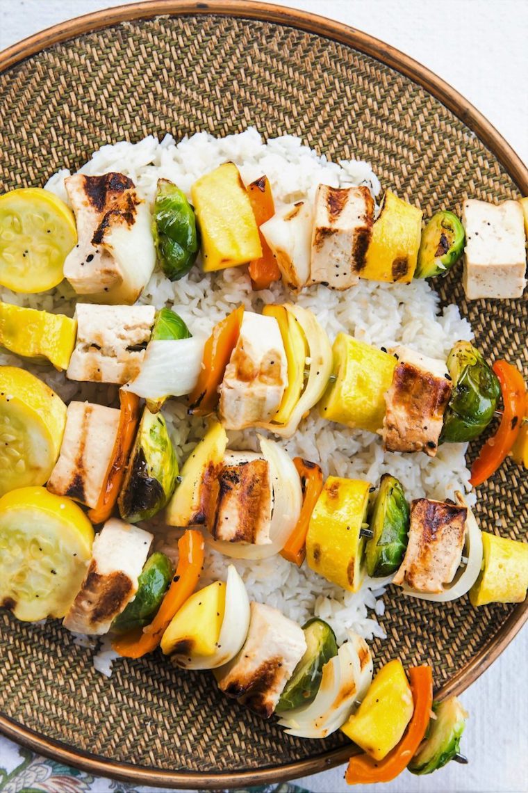 Birds eye view image of kabobs containing vegan sesame ginger tofu, zucchini, brussel sprouts, onions, and peppers, served on top of white, presented on a circular woven dish 