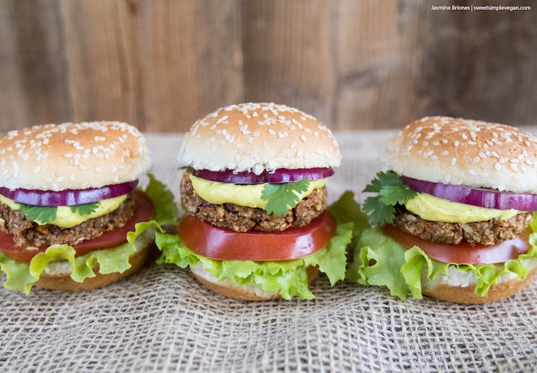 Medium long shot of three vegan veggie burger sliders inside sesame hamburger buns, garnished with lettuce, tomato, onion, fresh herbs, and a cashew mustard cheese sauce