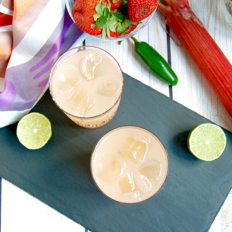 Birds eye view shot of two spicy strawberry margaritas alongside fresh lime, jalapeno peppers, and whole strawberries on a dark surface 