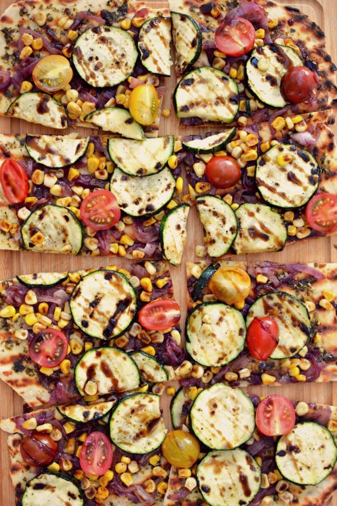 Birds eye view close up image of vegan veggie grilled square pizza slices containing pesto, zucchini, onions, tomatoes, and corn, garnished with balsamic, and served on a wooden platter