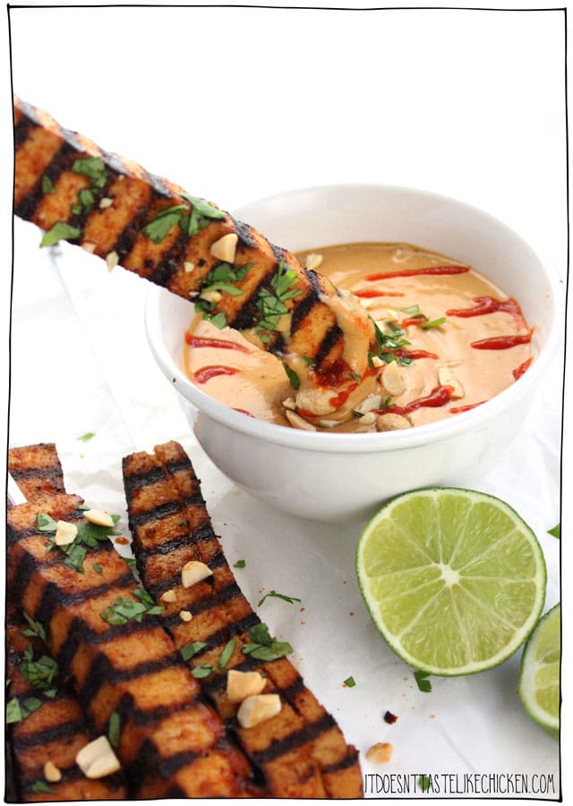 Close up image of a vegan grilled tofu skewer being dipped into a white bowl containing spicy peanut sauce alongside other tofu skewers, garnished with fresh lime, herbs, and chopped peanuts