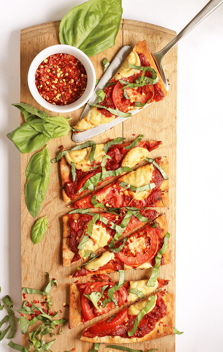 Birds eye view image of triangle slices of grilled vegan margherita pizza served on a rectangular wooden serving board, garnished with additional fresh basil and a small white ramekin of dried chilli flakes