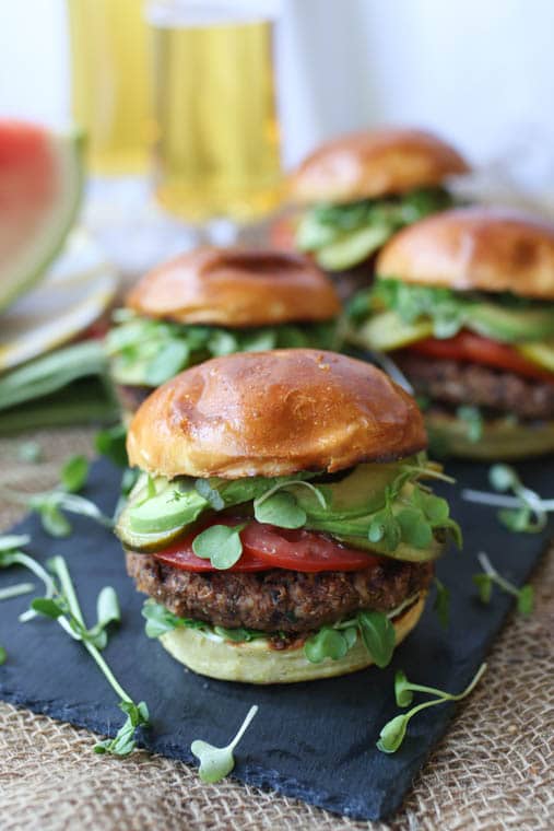 Close up of four vegan walnut and quinoa burgers served inside hamburger buns with avocado, pickles, tomatoes, and greens, on a long rectangular plate