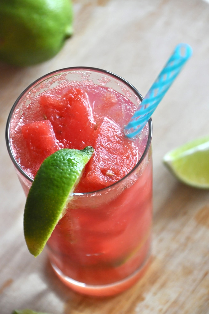 Close up of a vegan watermelon limemade cocktail presented in a large clear glass, served with a blue straw and a wedge of fresh lime