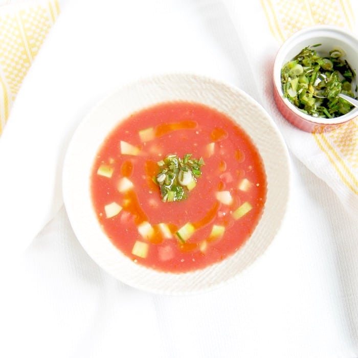 Birdseye view shot of vegan watermelon tomato gazpacho in a large white bowl garnished with scallion-cilantro relish, served next to a small red ramekin containing additional scallion-cilantro relish