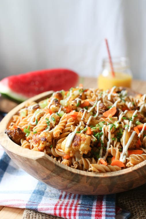 Close up of plant-based buffalo cauliflower pasta salad presented in a large wooden bowl, garnished with fresh chives, featuring a cocktail with a straw and a slice of watermelon in the background