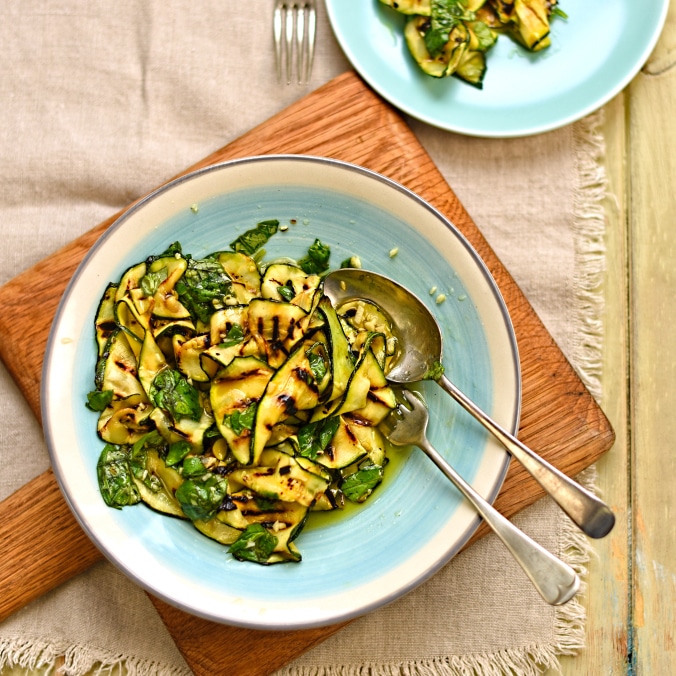 Birdseye view of chargrilled courgette salad with lemon and basil, served in a light blue bowl on top of a wooden serving plate and two silver serving utensils 