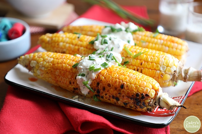 Close up image of three ears of corn on the cob garnished with vegan blue cheese dressing and chives, served on a square white serving dish with red detailing 