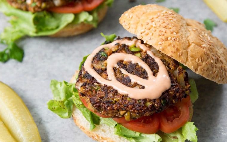 Close up of a vegan edamame and ancient grain burger served on a sesame whole grain bun with lettuce and tomatoes, garnished with aioli and a pickle on the side.