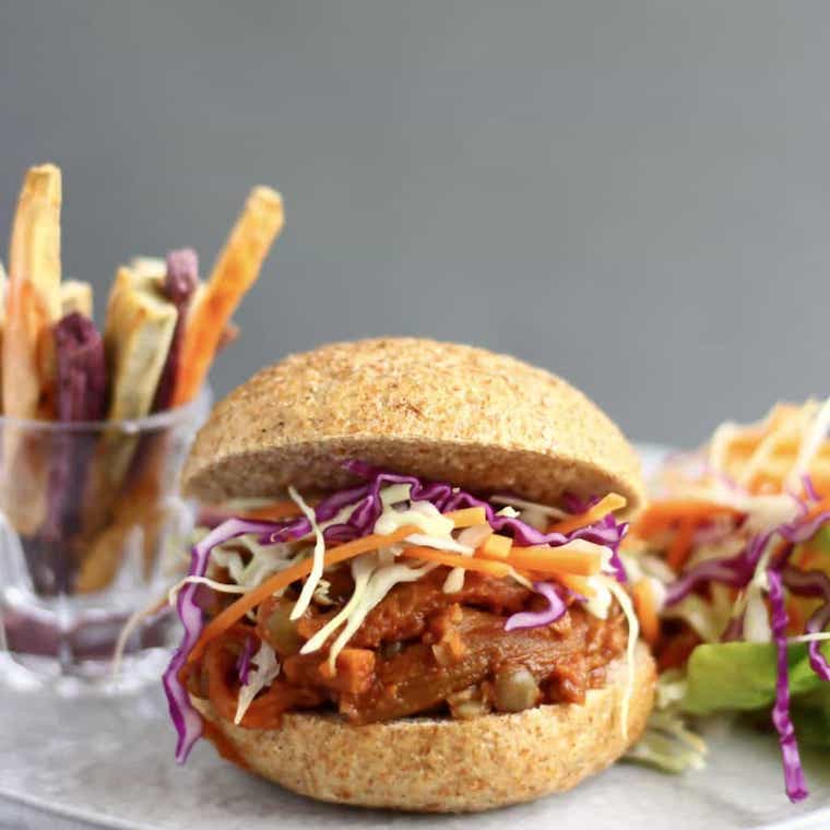 Close up image of plant-based eggplant pulled pork burger inside of a hamburger bun also containing coleslaw, with a clear glass in the background containing oven-baked fries of assorted colours 