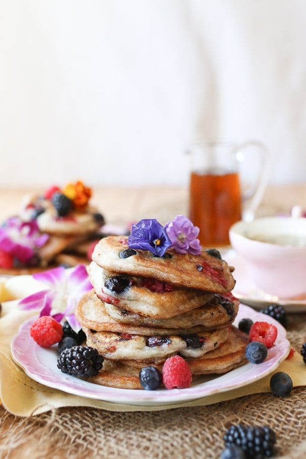 A pink plate with a stack of elderflower pancakes with berries scattered around.