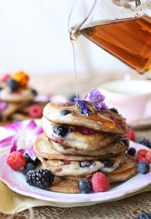 An angled image of a plate of elderflower pancakes with berries and edible flowers around with syrup poured on top.