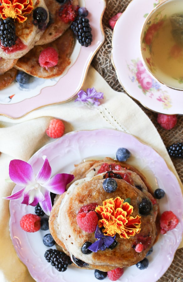 An overhead image of two plates of elderflower pancakes with berries and edible flowers around.