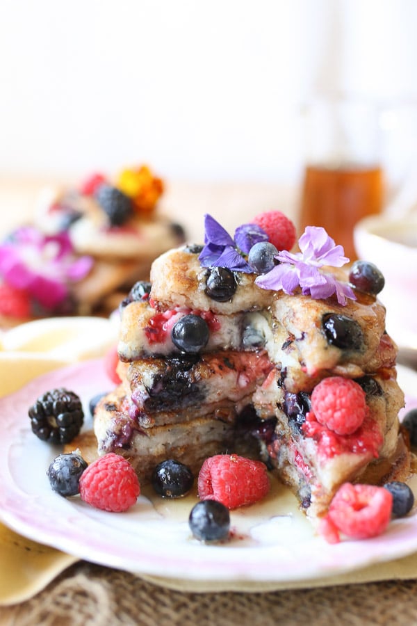 A side image of a plate of elderflower pancakes with berries and edible flowers around with a quarter cut opened.