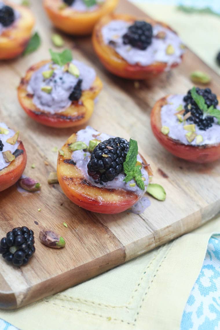 Close up image of several grilled peaches topped with blackberry coconut cream garnished with blackberries, pistachios, and mint, presented on a square wooden serving board