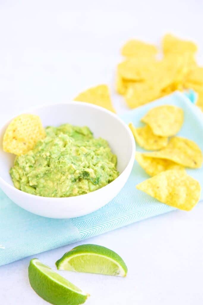 Full shot image of easy plant-based guacamole in a white bowl garnished with a corn chip, served with additional corn chips on the side atop of a blue surface